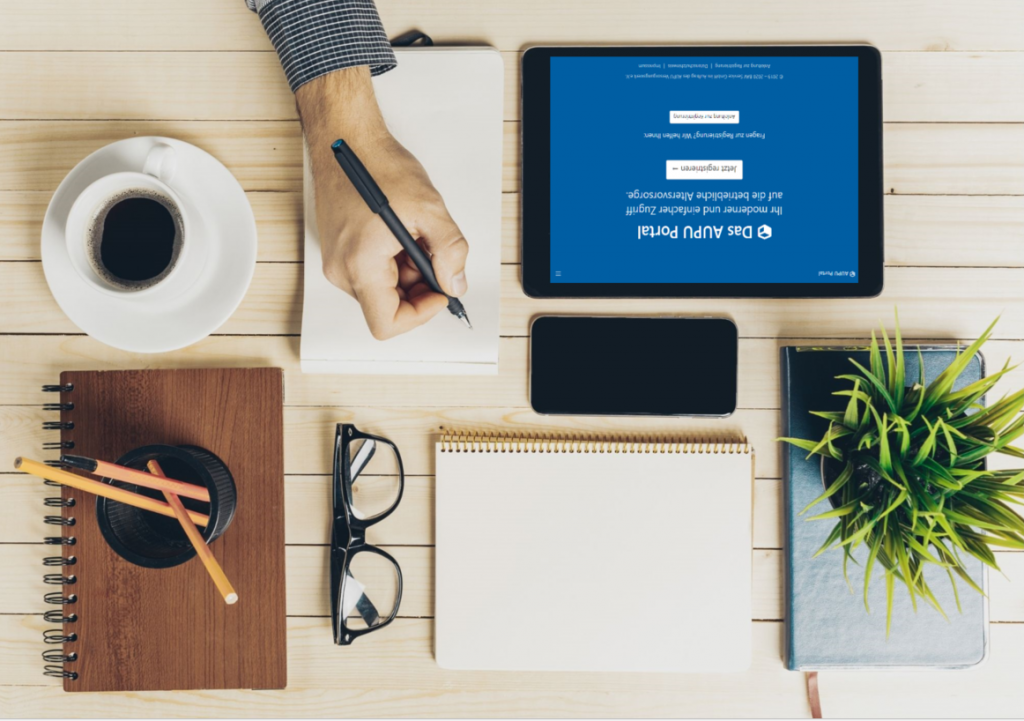 Oberansicht eines Schreibtischs mit Tablet, Smartphone, Notizbüchern, Stiften, einer Brille, einer Kaffeetasse und einer Pflanze. Eine Person schreibt in ein Notizbuch.