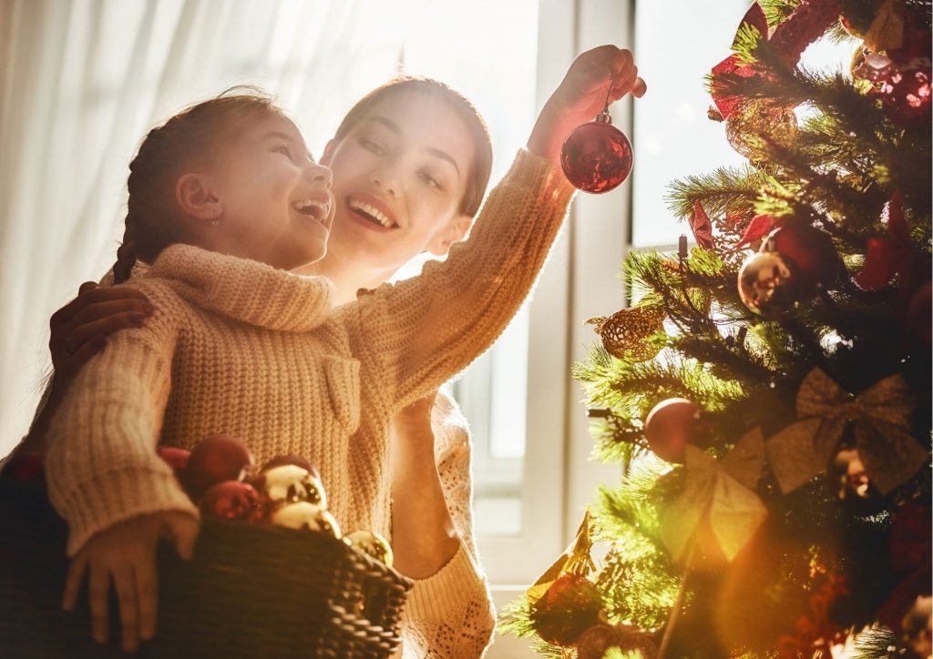 Eine lachende Mutter und ihre Tochter dekorieren einen Weihnachtsbaum mit roten Kugeln und goldenen Schleifen.